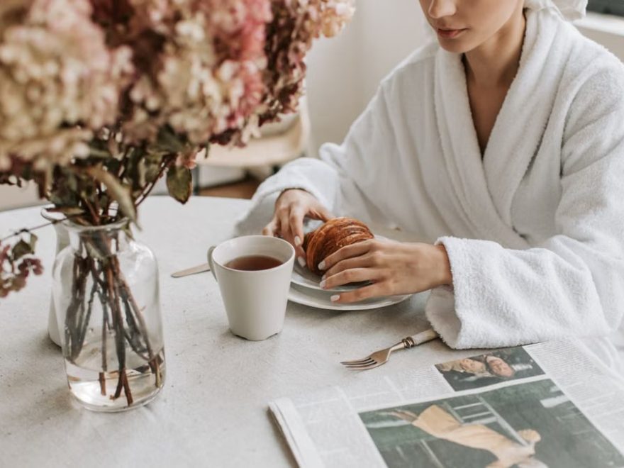 A woman in a bathrobe having breakfast indoors with a croissant and tea.